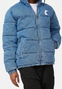 Denim puffer jacket in light blue with quilted texture, featuring a front zipper, side pockets, and elastic cuffs for optimal fit.
