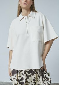 Polo-shirt i cremefarvet kortærmet ribbet stof med halv lynlås og en brystlomme. Skjorten har en afslappet pasform og rene linjer.