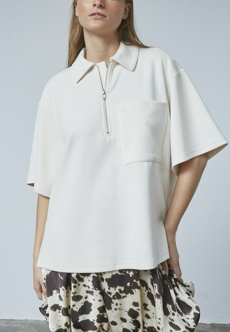 Polo-shirt i cremefarvet kortærmet ribbet stof med halv lynlås og en brystlomme. Skjorten har en afslappet pasform og rene linjer.
