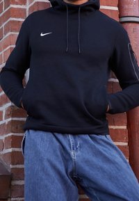 Felpa nera Nike con coulisse, maniche lunghe e tasca frontale. Abbinata a jeans in denim azzurri, sullo sfondo di mattoni.