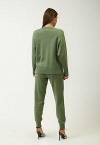 Groene gebreide trui met een ontspannen pasvorm en geribbelde zoom, gecombineerd met bijpassende joggers, beide met een gladde textuur en taps toelopende pijpen. Zilveren hakken zorgen voor contrast.