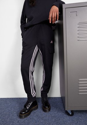 Osoba w czarnych spodniach dresowych Adidas z białymi paskami, czarnych butach i ciemnej bluzie opiera się o szarą metalową szafkę na niebieskim dywanie.