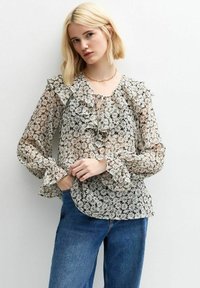 Blusa a maniche lunghe con rouches e motivo floreale in nero, bianco e verde. Presenta un scollo con lacci e tessuto strutturato. Abbinata a jeans blu.