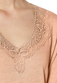 Pfirsichfarbener Oberteil mit tiefem V-Ausschnitt und filigranen Spitzenverzierungen entlang des Ausschnitts, das ein florales und schwirliges Muster hervorhebt.
