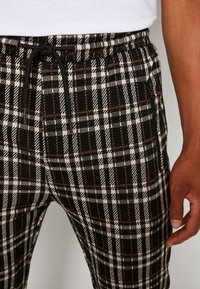 Svarta, vita och bruna rutiga byxor med dragsko i midjan och sidoficka med dragkedja, bärs av en person i en vit skjorta.