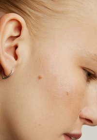 Pendiente plateado en forma de corazón colocado en la oreja, mostrando un acabado metálico suave. El tono de piel es claro, con textura y manchas visibles.