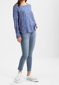 Blusa blue a maniche lunghe con motivo floreale, abbinata a jeans skinny in denim chiaro con decorazioni simil perla. Completato con sneakers bianche.