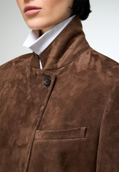 Közeli kép egy személyről, aki barna színű, magas galléros, velúr dzsekit visel fehér inggel alatta, a felső testére fókuszálva.