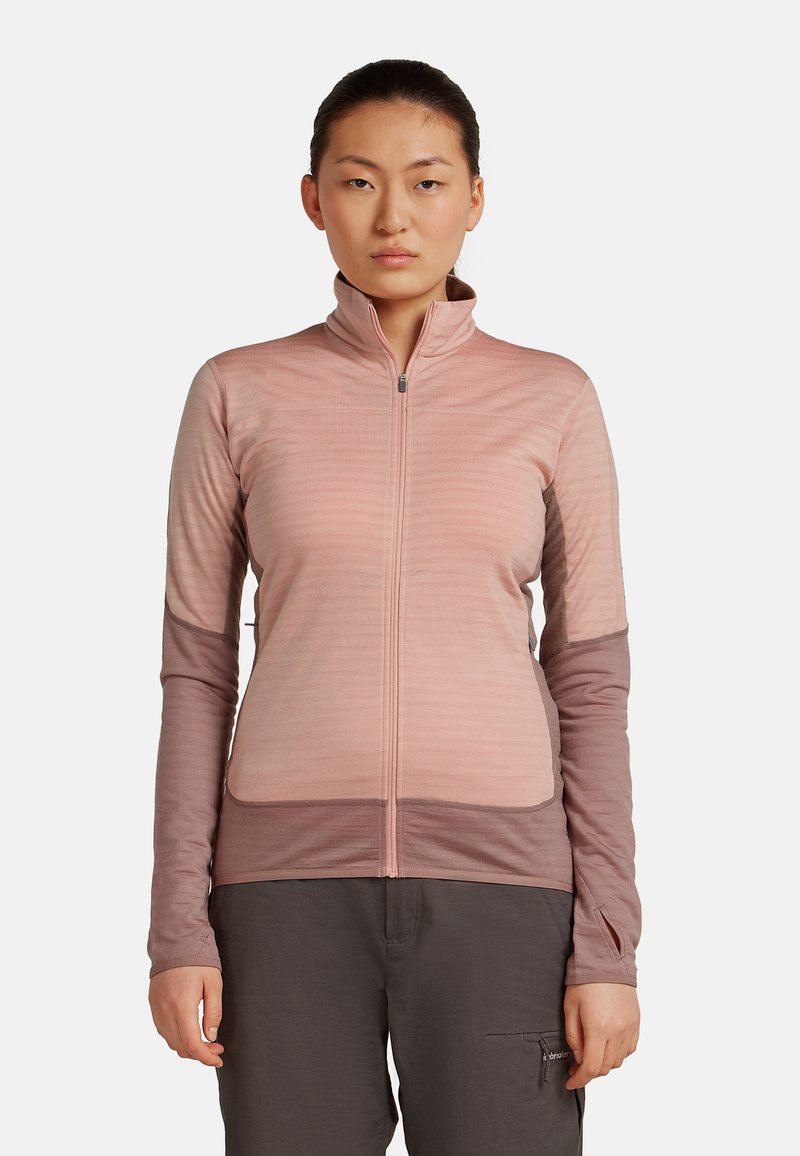 Mujer con el cabello oscuro recogido, vistiendo una chaqueta rosa con cremallera y pantalones oscuros, de pie contra un fondo blanco liso.