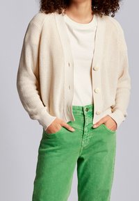 Ljusbeige stickad kavaj med knäppning framtill, ribbade ärmslut, bärs över en krämfärgad skjorta och gröna jeans, som visar en avslappnad passform och textur.