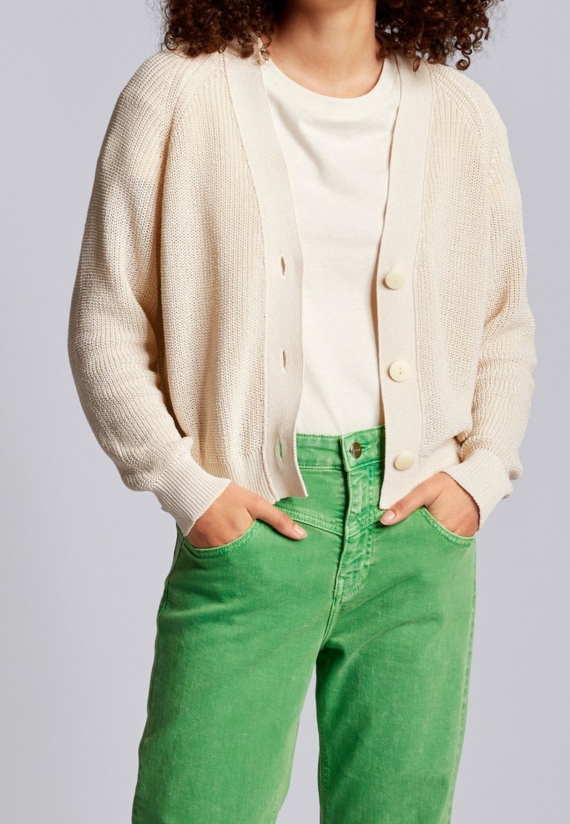 Ljusbeige stickad kavaj med knäppning framtill, ribbade ärmslut, bärs över en krämfärgad skjorta och gröna jeans, som visar en avslappnad passform och textur.