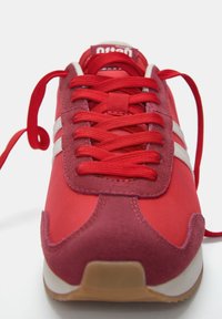 Baskets rouges présentant un dessus en textile lisse, des accents en daim, des rayures blanches et des lacets plats rouges. Semelle en caoutchouc avec un motif texturé.