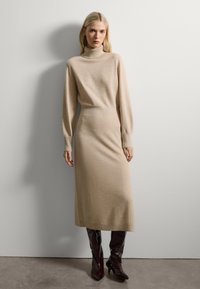 Vestido de punto beige con cuello alto, mangas largas y una silueta ajustada, combinado con botas brillantes de cuero marrón oscuro hasta la rodilla.