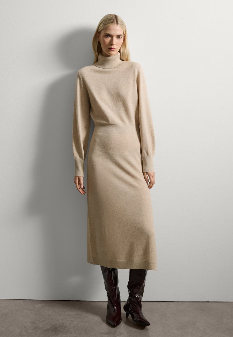 Vestido de punto beige con cuello alto, mangas largas y una silueta ajustada, combinado con botas brillantes de cuero marrón oscuro hasta la rodilla.