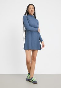 Sininen pitkähihaisen mekko, jossa on korkea kaulus, teksturoitu kangas ja levenevä hame. Yhdistettynä tummansinisiin kenkiin, joissa on neonvihreät nauhat.
