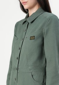 Veste en denim verte avec col, fermetures à boutons et une petite patch logo. Comprend des poches latérales et une coupe structurée.