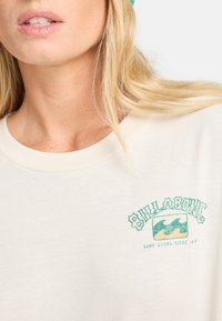 T-shirt w kolorze kremowym z zielonym logo z falami i tekstem. Klasyczny krój z okrągłym dekoltem, wykonany z miękkiego i gładkiego materiału.