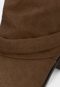 Brūna zamša potītes zābaki ar gludu tekstūru, salocītu dizaina detaļu un mazu papēdi. Apaļš purngalis ar elegantu siluetu.