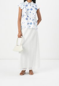 Donna che indossa una maxi gonna bianca in pizzo, una blusa corta a maniche corte blu e bianca con stampa di conchiglie, tacchi a strappi beige, mentre tiene in mano una piccola borsa bianca.