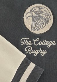 Ciemnoszara bluza z teksturowanym godłem orła i napisem "The College Rugby" w białym kursywie; kremowy rękaw z jasnymi paskami.