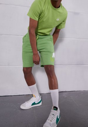 Persoon draagt een groene Nike T-shirt en korte broek, witte sokken en witte sneakers met een groene swoosh, staand tegen een lichtgrijze muur.