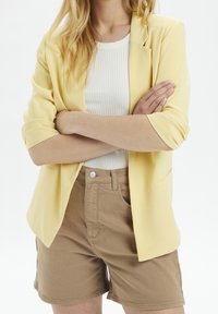 Yellow blazer with rolled sleeves, paired with a white ribbed shirt and beige shorts. Key features include a tailored fit and smooth texture.