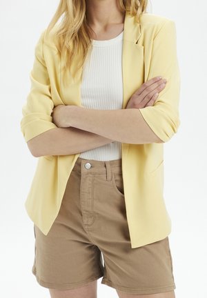 Blazer giallo con maniche arrotolate, abbinato a una maglietta bianca a coste e pantaloni beige. Le caratteristiche principali includono una vestibilità sartoriale e una trama liscia.