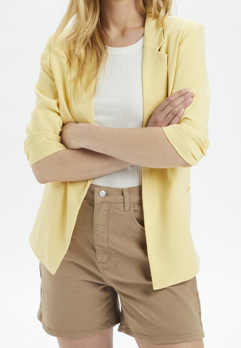 Yellow blazer with rolled sleeves, paired with a white ribbed shirt and beige shorts. Key features include a tailored fit and smooth texture.