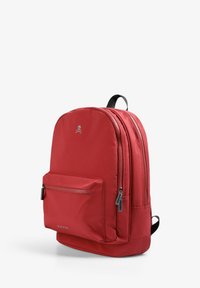 Mochila de nailon roja con textura suave, forma redondeada, dobles cremalleras, bolsillo frontal y acento de logo en la parte superior. Correas ajustables negras.
