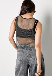 Top canottiera in mesh nero con design senza maniche, caratterizzata da una schiena semi-trasparente. Abbinata a pantaloni di jeans grigi a vita alta con tasche visibili.
