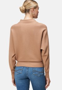 Pull en maille côtelée marron avec un col haut et des épaules tombantes, associé à un jean bleu avec un design décoratif sur la poche arrière.