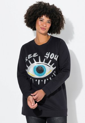 Zwarte sweatshirt met een grote oogafbeelding versierd met pailletten in blauw, zilver en zwart. Boven het oog staat de tekst "SEE YOU". Lange mouwen.