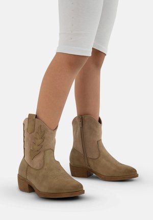 Botas de vaquero de ante en tono marrón con costuras decorativas, cierre lateral de cremallera y tacón cuadrado, mostradas en pies con leggings blancos.