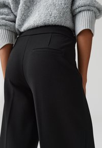 Zwarte broek met een gladde textuur, voorzien van een achterzak, gedragen met een grijze, gestructureerde trui met ribbelmanchetten.