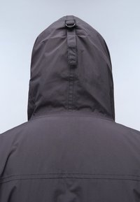 Grå vandtæt jakke med hætte, tekstureret stof og en løkke bagpå. Enkel design med synlige sømme langs ryggen.