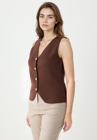 Kobieta z jasnobrązowymi włosami nosi bezrękawnik w kolorze brązowym z guzikami oraz beżowe spodnie, stojąc na tle jednolitego białego tła.