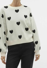 Suéter de crema claro con patrones de corazones negros, textura de punto suave, mangas anchas y puños acanalados. Combinado con jeans negros.