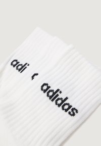 Para białych skarpetek typu crew z żeberkami, z czarnym haftowanym logo Adidas na mankiecie, wyłożona na jasnym tle.
