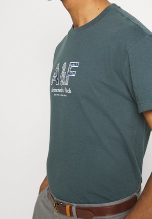 Mężczyzna w ciemnozielonym t-shircie Abercrombie & Fitch wpuszczonym do szarych spodni z brązowym paskiem na jednolitym tle.
