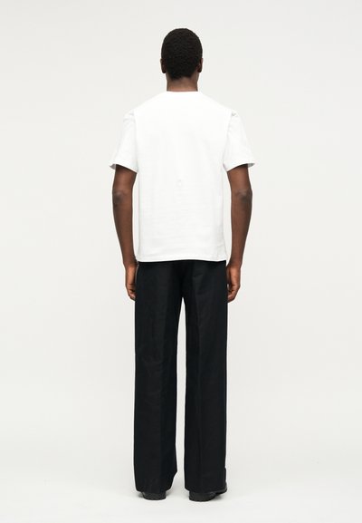 Άνδρας που στέκεται με πλάτη, φοράει απλό λευκό κοντομάνικο πουκάμισο και φαρδιά μαύρα παντελόνια σε ένα μονόχρωμο φωτεινό φόντο.