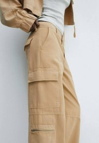 Beige cargo bukser lavet af letvægtsstof, med flere lommer, en lynlåsdetaile, og et afslappet fit design.