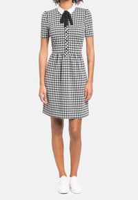 Musta-valkoinen ruudullinen mekko, jossa on kauluspanta ja nauhakiinnitys, lyhyet hihat, napitettava etuosa ja liehuva helma. Valkoiset tennarit.