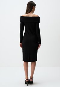 Abito a maglia nero, scollato sulle spalle, con maniche lunghe, silhouette aderente, taglio a lunghezza ginocchio e dettaglio dello spacco sul retro. Texture liscia, design minimalista.