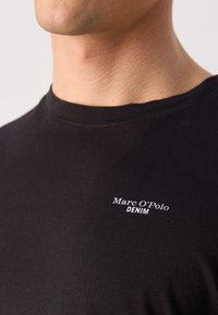 Tricou din bumbac negru, cu guler rotund și un logo imprimat alb „Marc O'Polo DENIM” în partea stângă a pieptului.