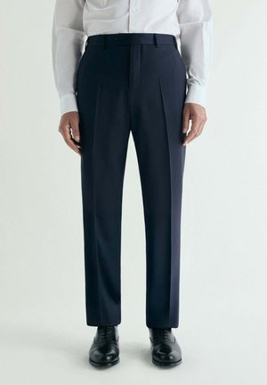 Hombre con pantalones de vestir azul marino a medida, camisa blanca con botones y zapatos de vestir negros pulidos, de pie frente a un fondo liso.