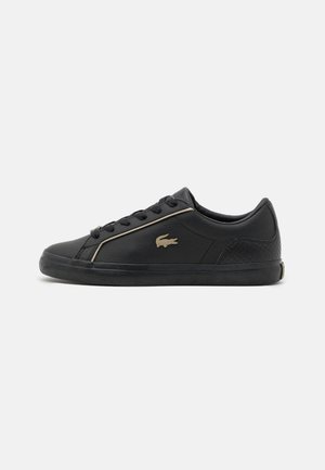 Czarny niski sneaker z czarnymi sznurowadłami, złotymi zdobieniami i złotym logo krokodyla po boku, płaska gumowa podeszwa, wszystko na prostym tle.