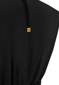 Vêtement noir avec une texture lisse, ornée d'une étiquette en métal doré avec des détails gravés, conçu avec une coupe ample et des plis subtils.