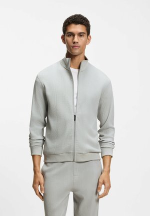 Ung mand iført lys grå tekstureret lynlåstrøje og matchende bukser, stående foran en ensfarvet hvid baggrund.