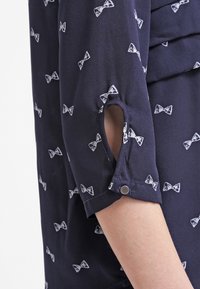 Blouse bleu marine avec imprimé de nœuds blancs, manches trois-quarts avec détail de boutons, tissu doux et finition légèrement texturée.