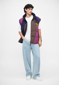 Colete acolchoado multicolorido com secções roxas, castanhas e verdes, usado por cima de uma camisa branca. Combinado com jeans de perna larga em azul claro e ténis brancos.